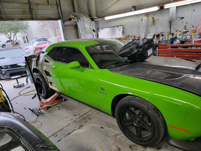 A lime green Dodge Challenger with a dark hood inside a busy repair shop with other vehicles