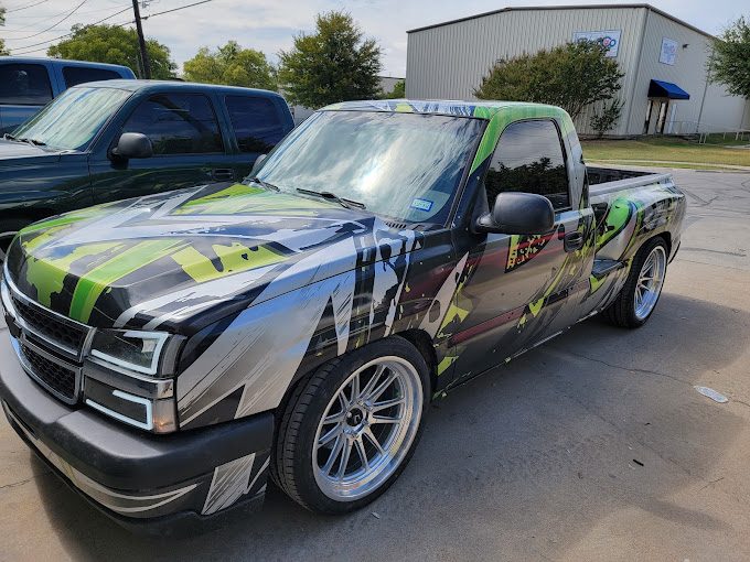 A wrapped Chevrolet Silverado pickup with black, green, and white graphics beside a metal building