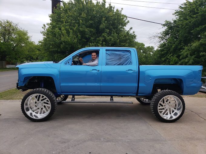 A lifted bright blue Chevrolet pickup with polished wheels, the driver waving from the window