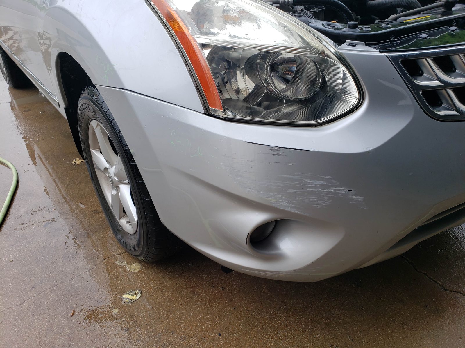 Close-up of the scuffed front bumper and fog light area of a silver Nissan with its hood open
