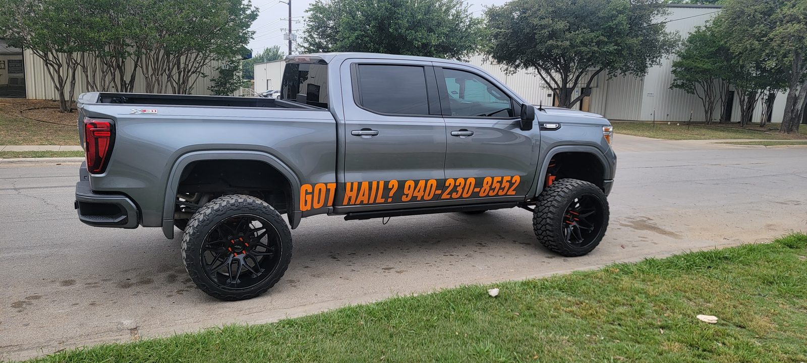 A gray GMC Sierra pickup with Got Hail lettering on the door, parked on grass beside a road